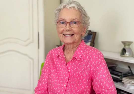 Elderly woman with short white hair, wearing glasses and a bright pink floral shirt, smiles warmly while seated in a cozy, light-filled room.